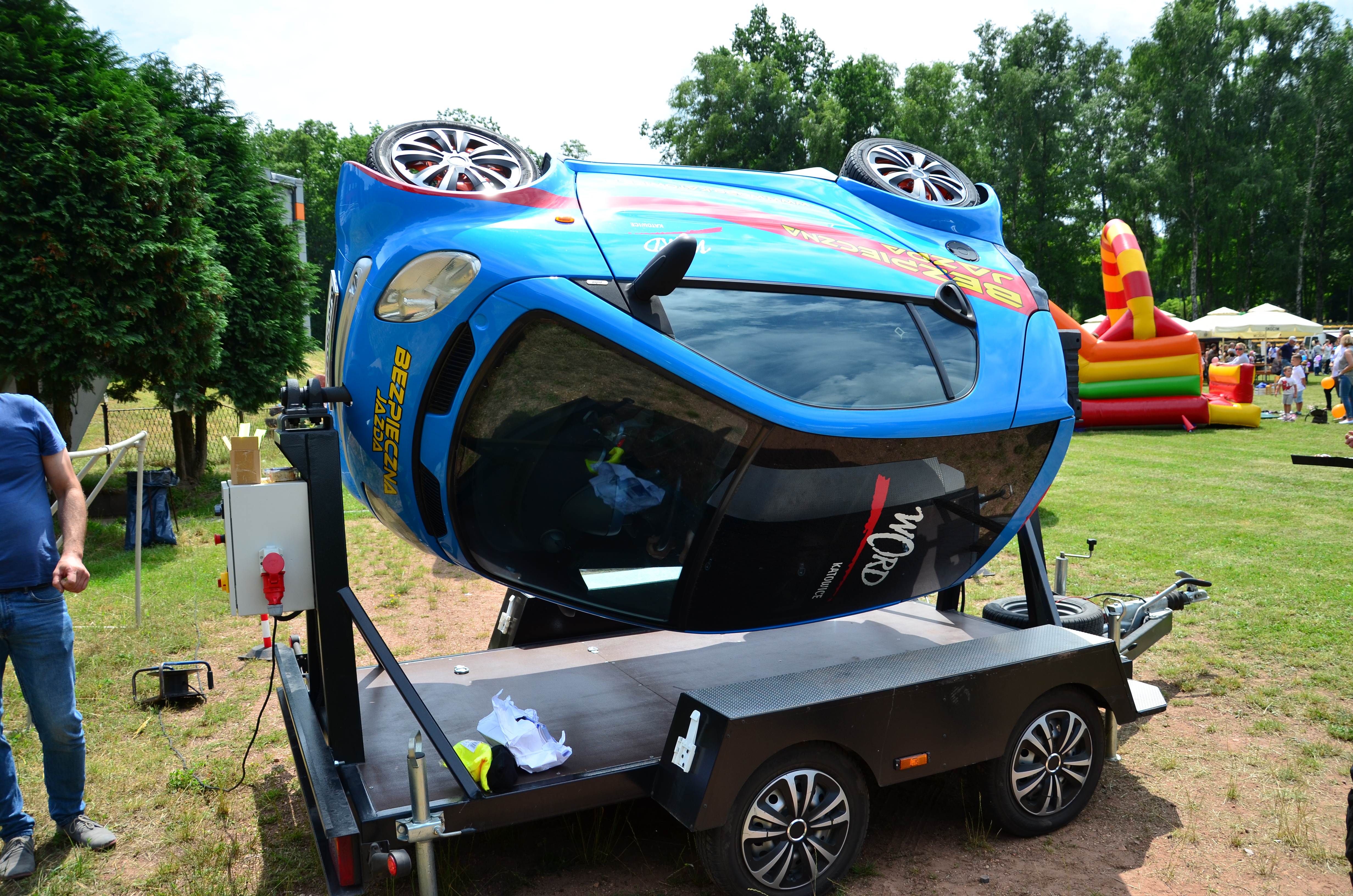 Zdjęcie przedstawia symulator dachowania samochodu. Niebieski samochód na przyczepie