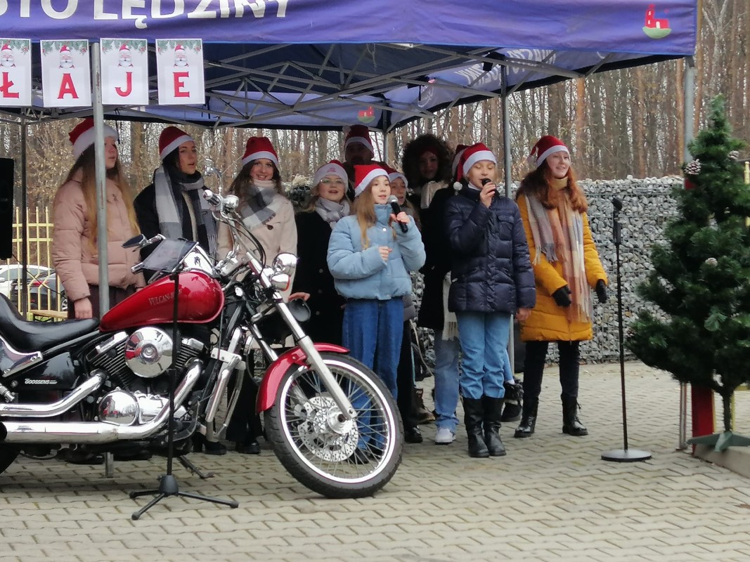 10 dziewczyn wykonuje piosenkę. Stoją i śpiewają. Przed nimi stoi zaparkowany czerwony motocykl