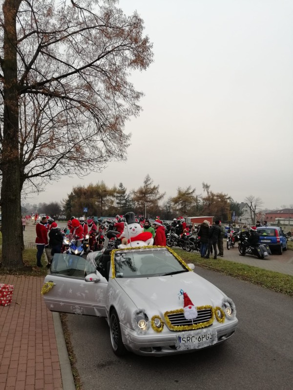 srebrny kabriolet mercesed zaparkowany na ulicy. Samochód przystrojony swiątecznymi elementami. Z tyłu motocykliści przebrani za mikołajów
