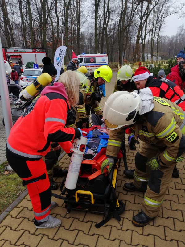 ratownicy medyczni oraz strażacy wykonują pokaz ratownictwa. pomiędzy nimi leży osoba, na której pokazują przebieg akcji
