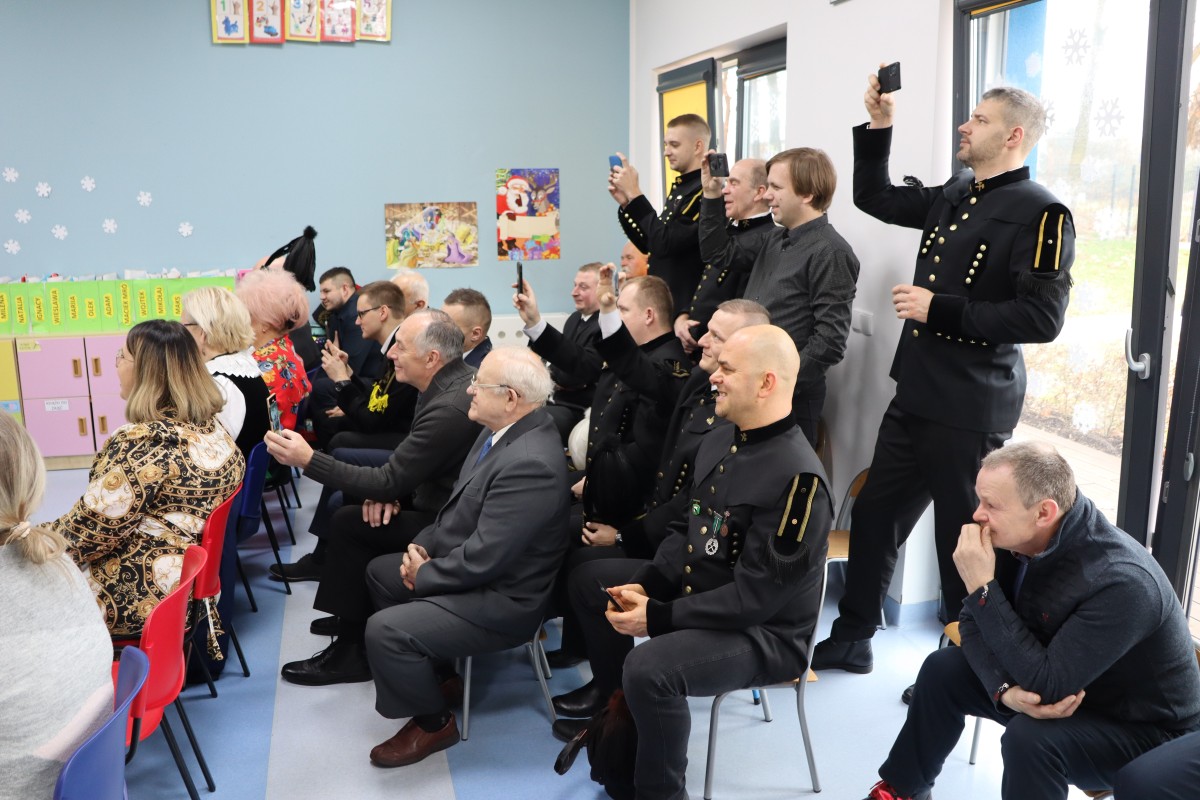 Ojcowie i dziadkowie ogladają występy dzieci. Część mężczyzn jest w strojach góniczych. Kilka osób nagrywa występy telefonami