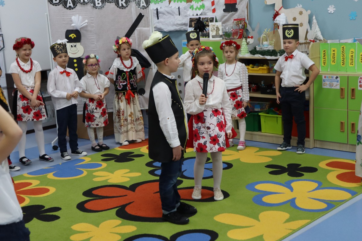 Dziewczynka stoi z chłopcem na środku dywanu, mówi do mikrofonu. Wokół stoją pozostałe dzieci