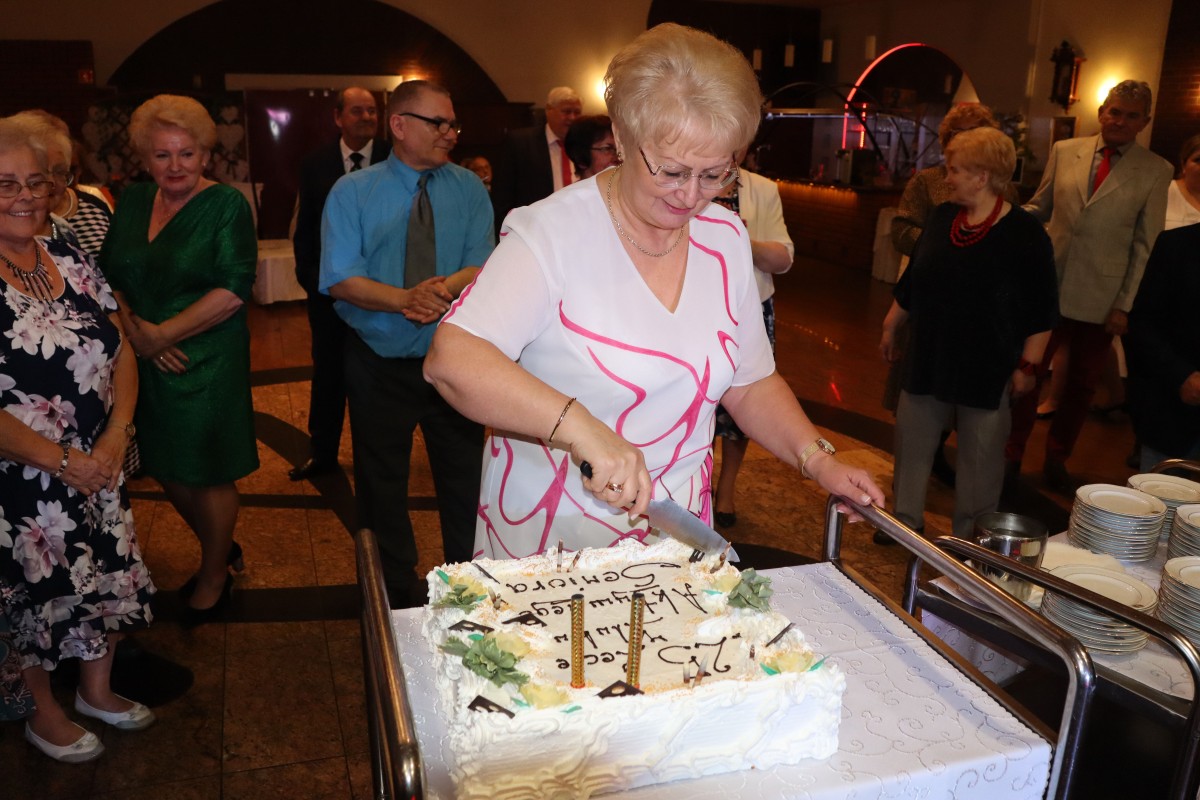 kobieta kroi tort, z tyłu obserwują to inni ludzie