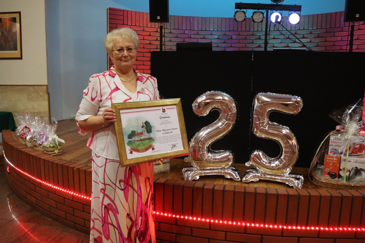 Kobieta pozuje trzymając w rękach dedykację w złotej ramie. za nią napis 25 z balonó i kosze prezentowe