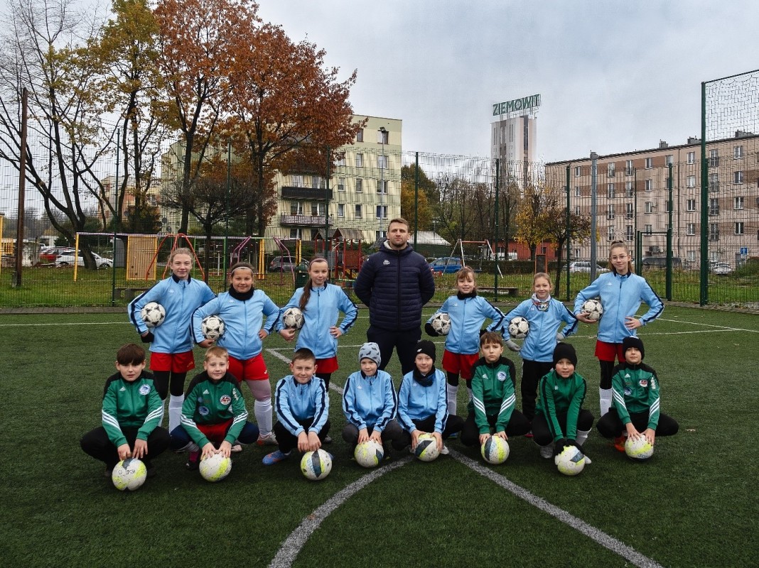 Dzieci w strojach piłkarskich pozują do zdjęciach. Na środku trener