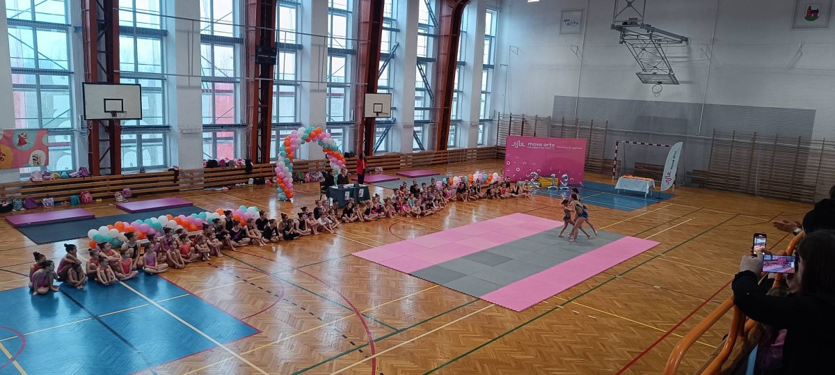 zawodniczki w sportowych strojach prezentują układ akrobatyczny na różowo-szarej macie. Wokół nich siedzą na podłodze inne uczestniczki wydarzenia, ubrane w czarne i różowe stroje. Na trybunach widzowie robią zdjęcia i nagrywają występ na telefonach.