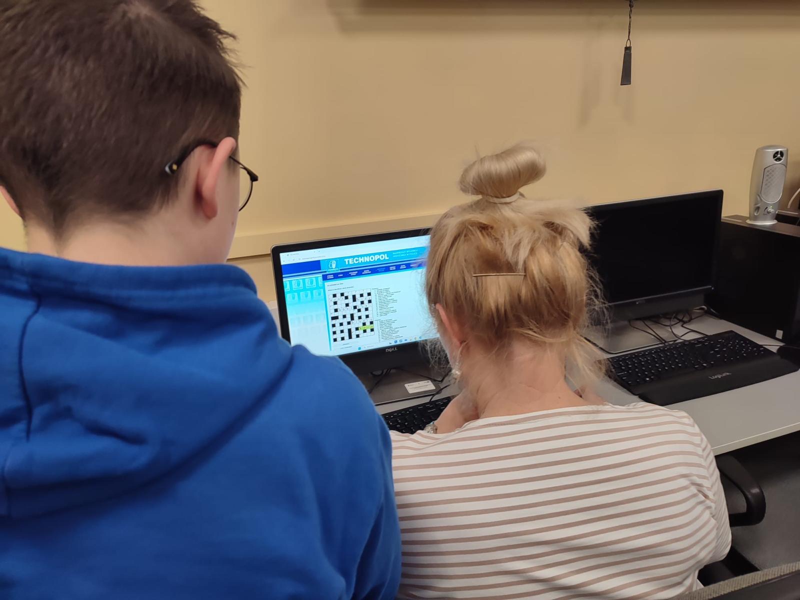 Seniorka podczas pracy przy komputerze, za nią uczeń pomagający jej.